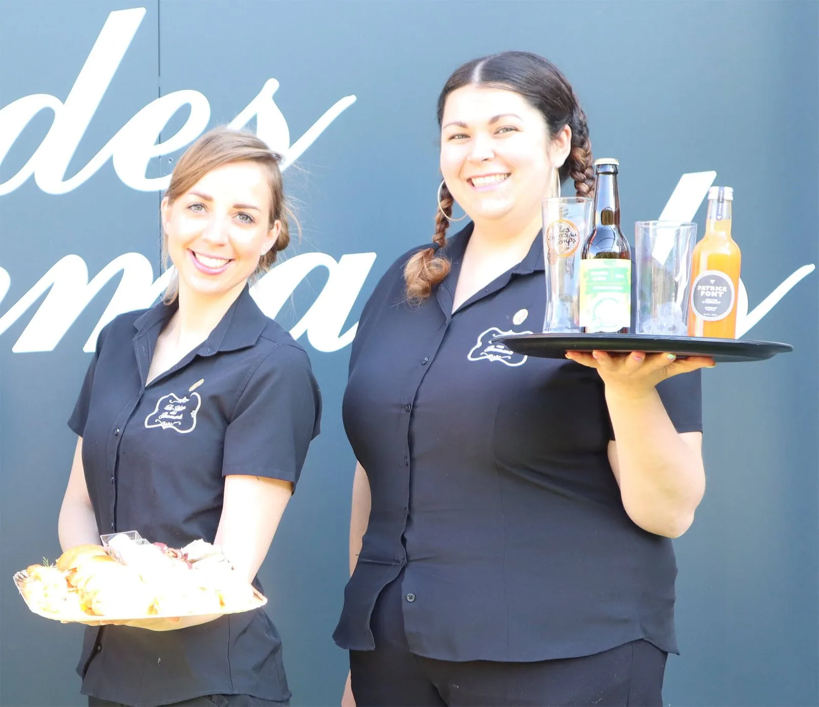 Deux membres de l'équipe de salle, souriantes, un plateau de bouchées et des boissons à la main
