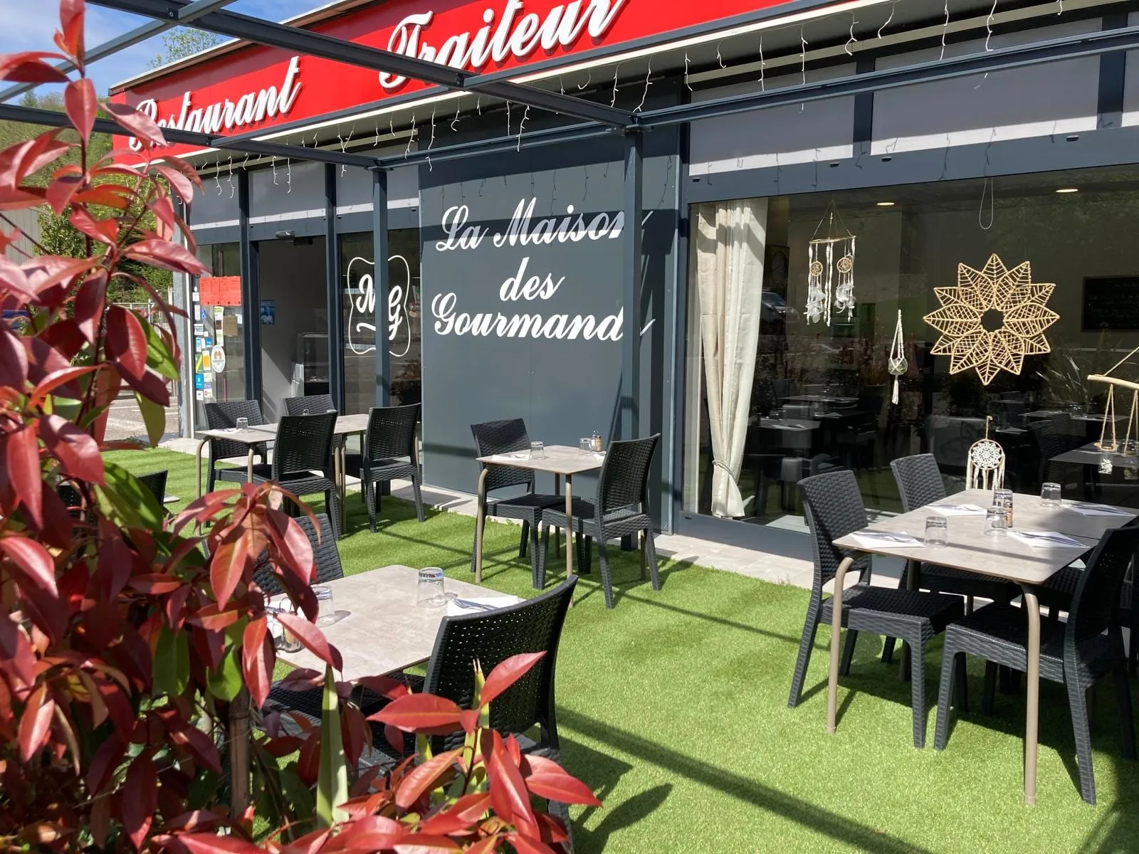 La terrasse du restaurant au soleil : tables dressées sur la pelouse, devant l'enseigne et le photinia