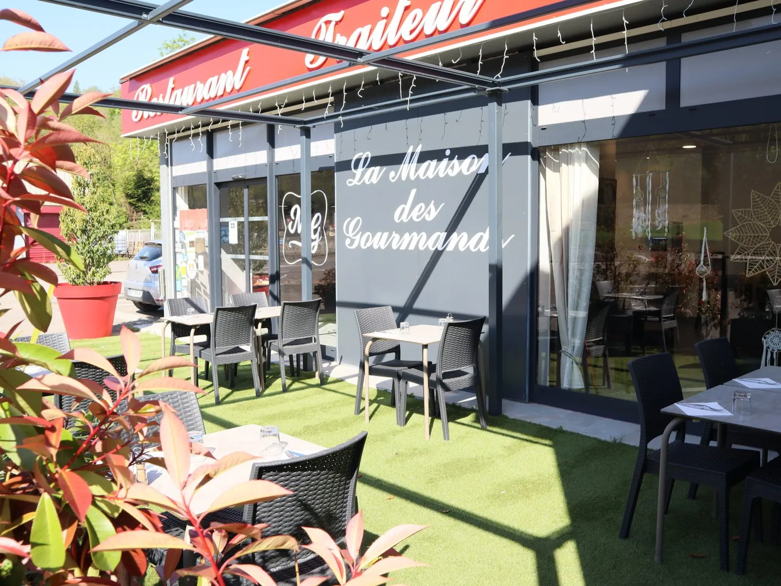 La façade du restaurant et sa terrasse : enseigne rouge Restaurant Traiteur, tables sur la pelouse au soleil