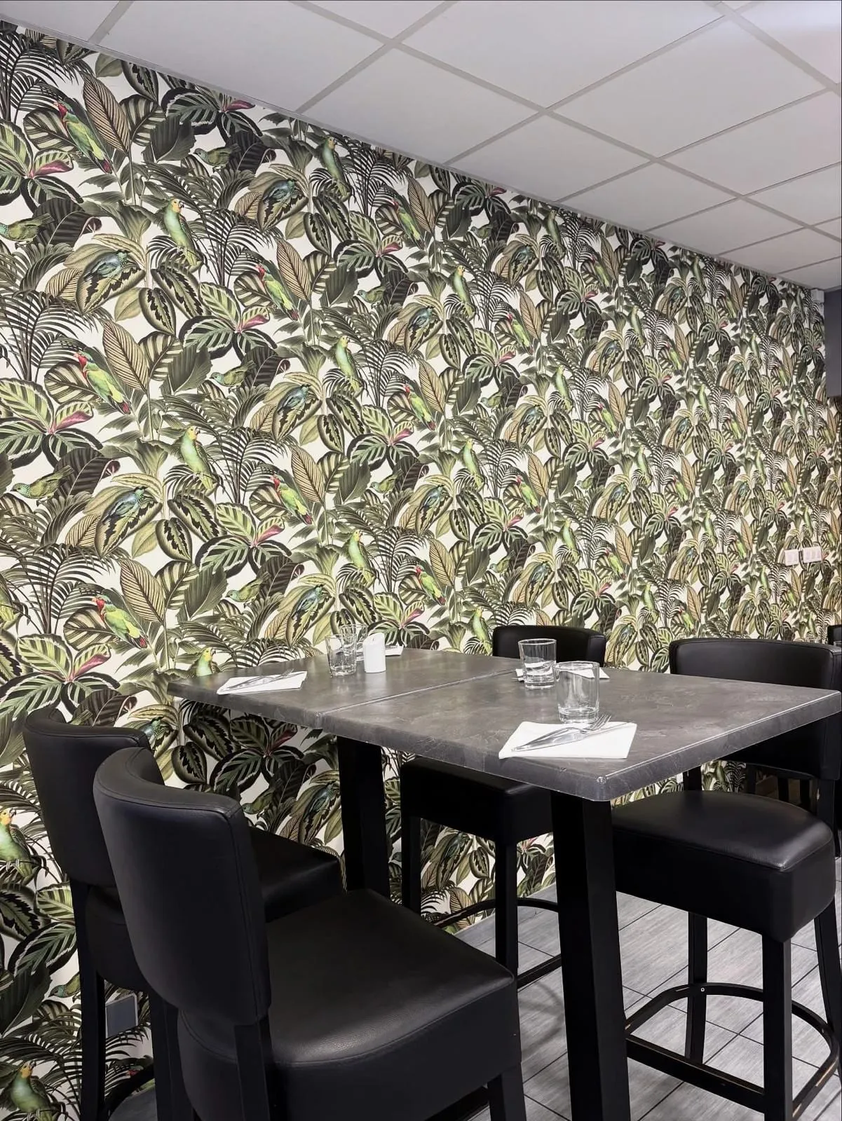 Un coin de la salle au papier peint tropical, table haute et tabourets noirs