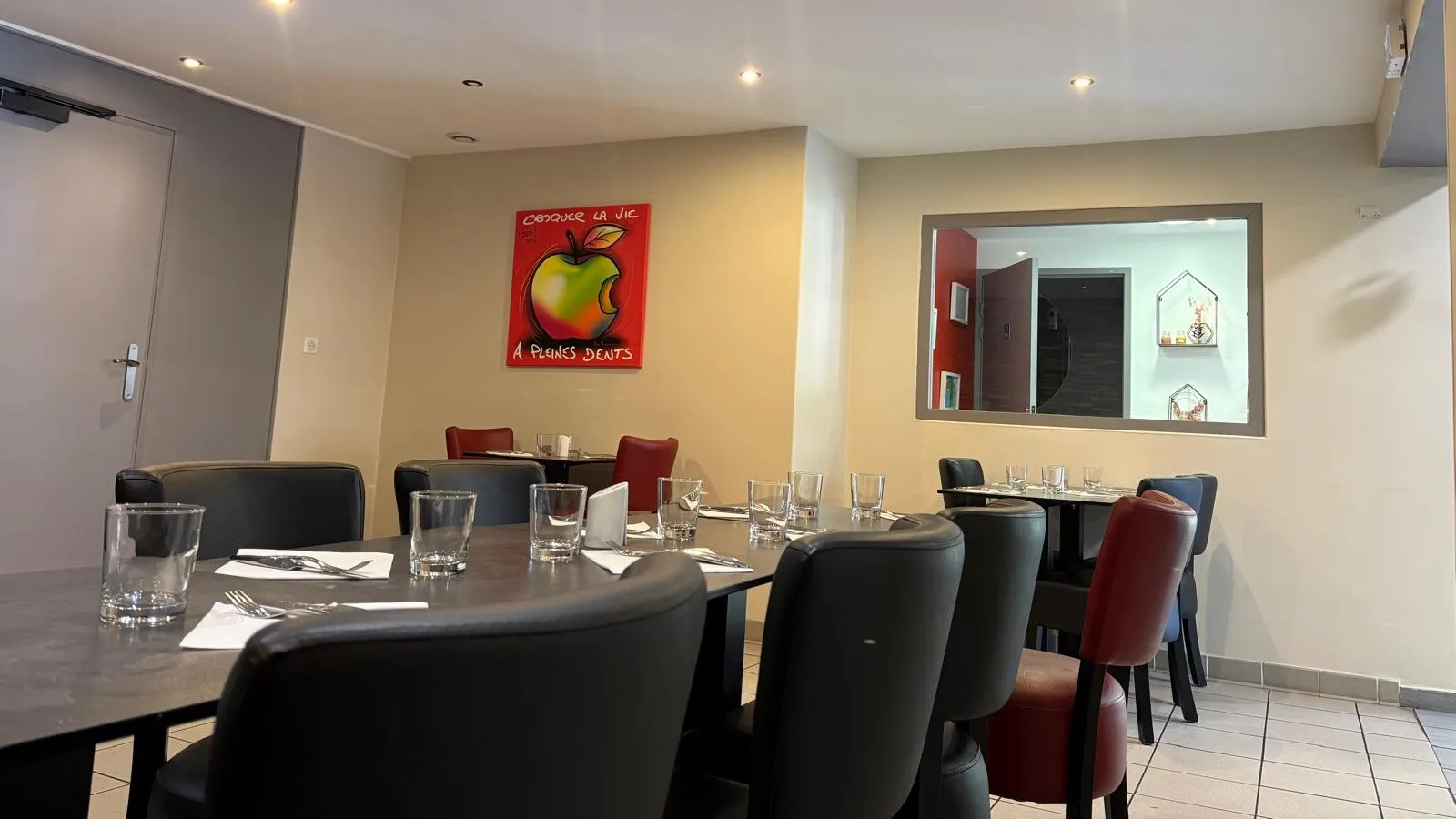 La salle du restaurant : tables dressées, fauteuils noirs et rouges, tableau « Croquer la vie à pleines dents »