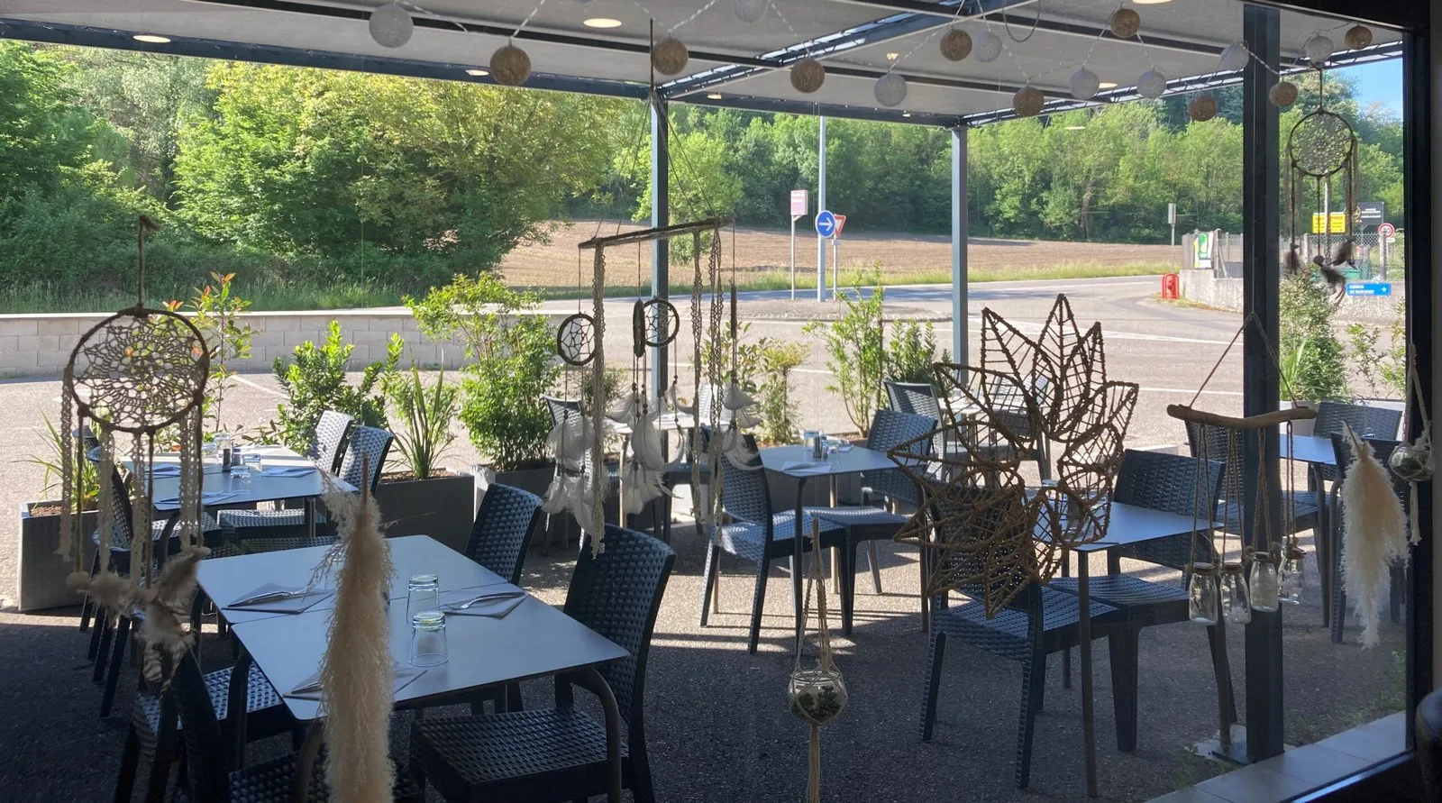 La terrasse couverte, tables dressées sous la pergola, attrape-rêves et verdure en arrière-plan